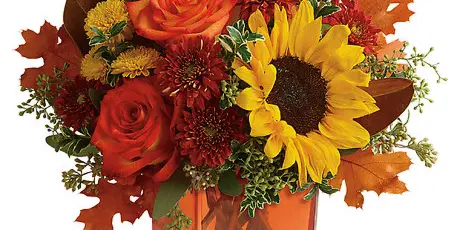 Autumn floral arrangement with red roses, sunflowers, and fall leaves in a square glass vase