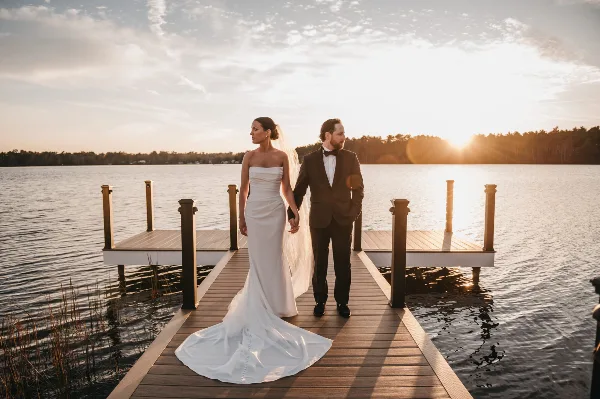 A bride and groom standing on a wooden dock by a lake at sunset.