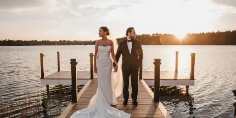 A bride and groom standing on a wooden dock by a lake at sunset.