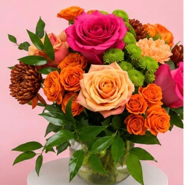 A colorful bouquet of pink, orange, and peach roses with green filler flowers arranged in a clear glass vase.