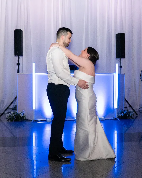 A bride and groom dancing in front of a DJ booth featuring professional speakers and vertical LED light bars.
