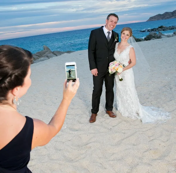 A bride and groom standing on a beach holding a bouquet while a photographer takes their picture with a smartphone.