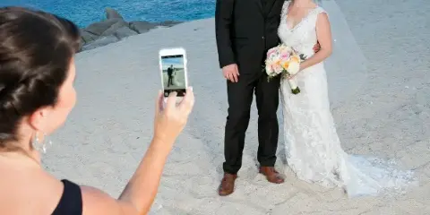 A bride and groom standing on a beach holding a bouquet while a photographer takes their picture with a smartphone.