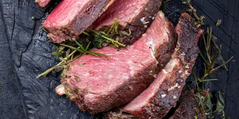Sliced cooked steak garnished with fresh herbs on a dark wooden serving board.
