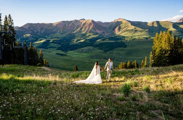 A bride and groom holding hands in a grassy field with a backdrop of green mountains and a ski lift.