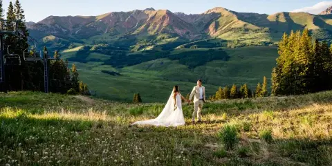 A bride and groom holding hands in a grassy field with a backdrop of green mountains and a ski lift.