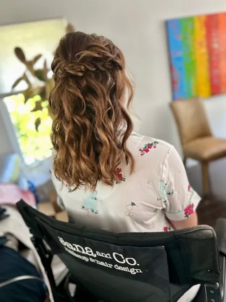 Back view of a woman with a styled wavy bridal updo sitting in a chair branded with the Jana and Co. makeup and hair design logo.