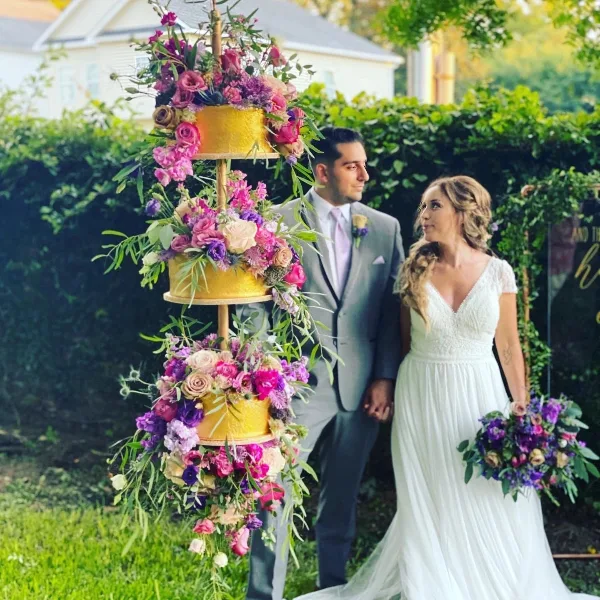 A three-tiered gold wedding cake decorated with vibrant pink and purple flowers, standing in a garden with a bride and groom in the background.