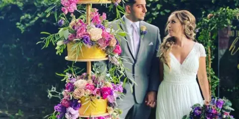A three-tiered gold wedding cake decorated with vibrant pink and purple flowers, standing in a garden with a bride and groom in the background.