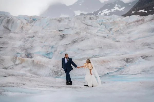 A bride and groom holding hands on a glacier with mountains in the background.