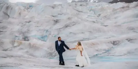 A bride and groom holding hands on a glacier with mountains in the background.