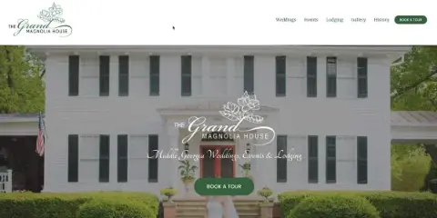 Exterior view of a white two-story historic antebellum house with dark shutters, a front veranda, manicured hedges, and a bride standing on the steps.