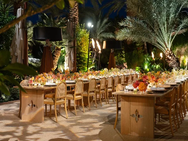 An outdoor wedding reception setup featuring long wooden farm tables and rattan-backed chairs arranged under palm trees at night.