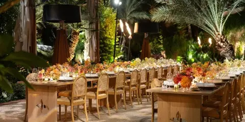 An outdoor wedding reception setup featuring long wooden farm tables and rattan-backed chairs arranged under palm trees at night.