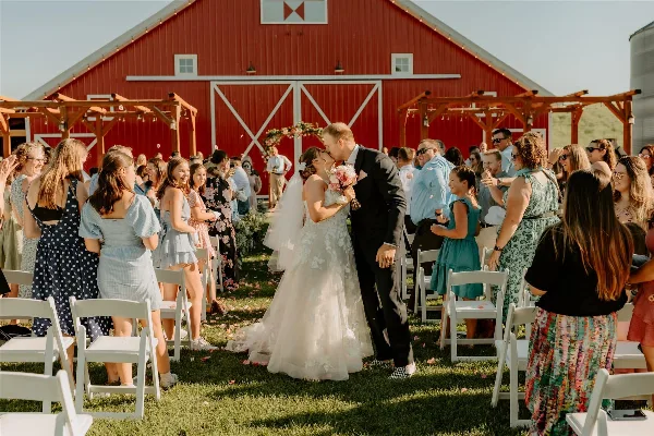 A bride and groom sharing a kiss during an outdoor wedding ceremony in front of a large red barn with white trim.