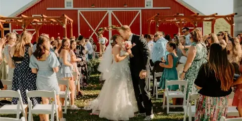 A bride and groom sharing a kiss during an outdoor wedding ceremony in front of a large red barn with white trim.