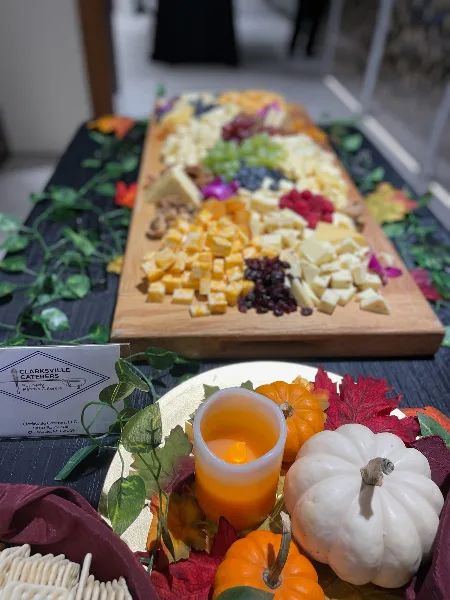 A catering table display featuring a wooden board with assorted cheeses, grapes, and dried fruit, accompanied by decorative pumpkins and a Clarksville Caterers business card.