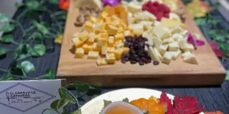 A catering table display featuring a wooden board with assorted cheeses, grapes, and dried fruit, accompanied by decorative pumpkins and a Clarksville Caterers business card.