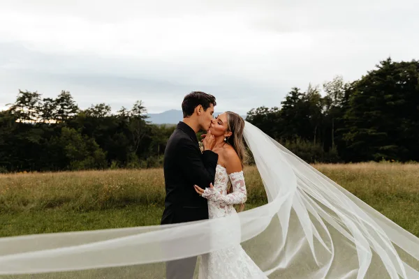 A bride and groom sharing a kiss in a grassy field with a long veil blowing in the wind.