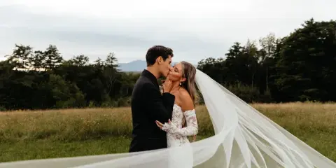 A bride and groom sharing a kiss in a grassy field with a long veil blowing in the wind.