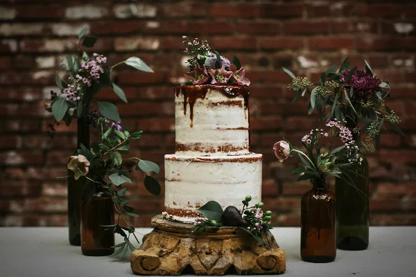 A two-tier semi-naked wedding cake decorated with fresh flowers, displayed on a wooden stand against a brick wall.