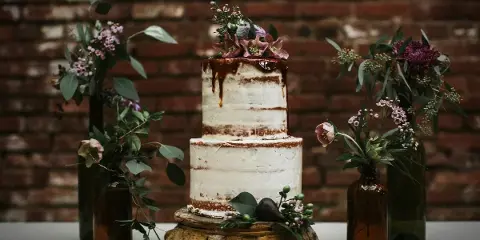 A two-tier semi-naked wedding cake decorated with fresh flowers, displayed on a wooden stand against a brick wall.
