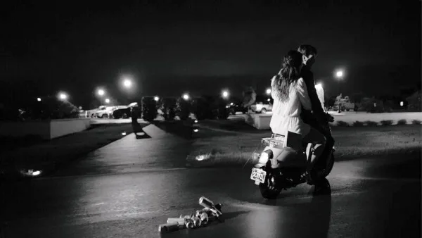 A black-and-white cinematic photograph of a couple sitting on a scooter at night in a parking lot.