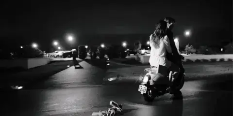 A black-and-white cinematic photograph of a couple sitting on a scooter at night in a parking lot.