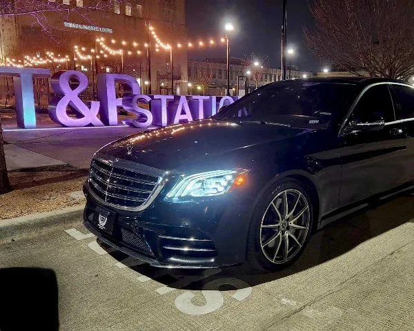 Black Mercedes-Benz S-Class sedan parked on a street at night with illuminated string lights in the background.