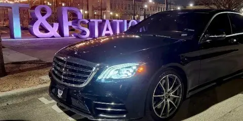 Black Mercedes-Benz S-Class sedan parked on a street at night with illuminated string lights in the background.