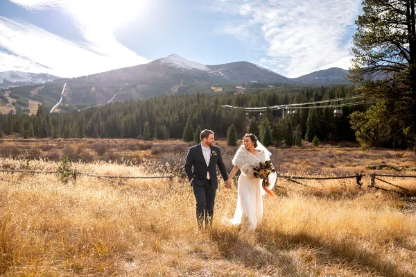A bride and groom walking hand-in-hand through a golden grassy field with a mountain and ski lift in the background.