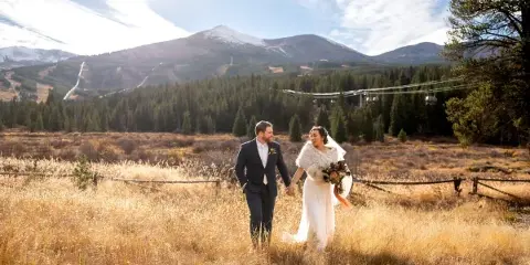 A bride and groom walking hand-in-hand through a golden grassy field with a mountain and ski lift in the background.