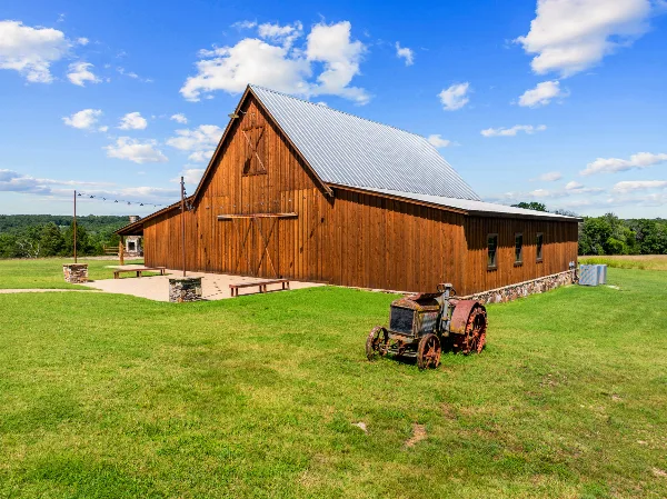A large wooden barn with a metal roof and outdoor stone fireplace situated on a grassy field under a blue sky.