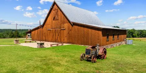 A large wooden barn with a metal roof and outdoor stone fireplace situated on a grassy field under a blue sky.