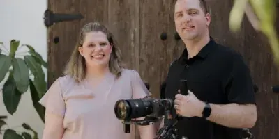 A man and a woman standing together holding professional video cameras and a gimbal stabilizer.
