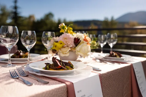 A plated dish of meat and vegetables on a white plate, set on a table with wine glasses and a floral arrangement.