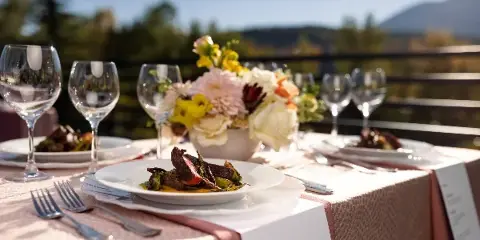 A plated dish of meat and vegetables on a white plate, set on a table with wine glasses and a floral arrangement.