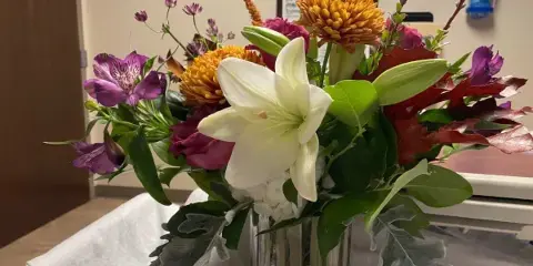 A vibrant floral arrangement with white lilies, purple alstroemeria, and orange chrysanthemums in a clear glass vase.