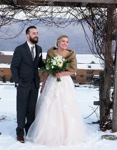A bride and groom standing together in a snowy landscape with a wooden structure and mountains in the background.