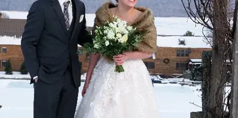 A bride and groom standing together in a snowy landscape with a wooden structure and mountains in the background.