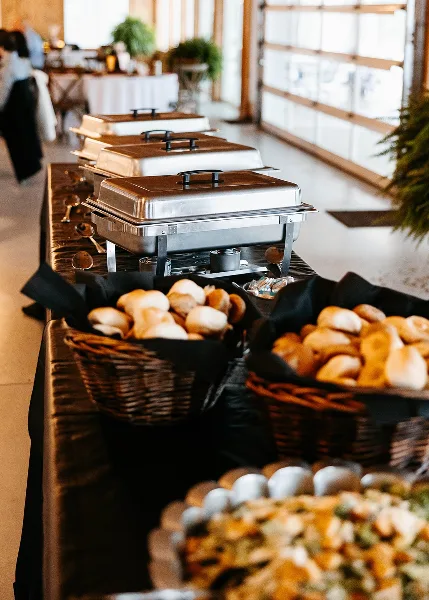 A catering buffet line with stainless steel chafing dishes and baskets of bread rolls set up in an event venue.