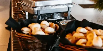 A catering buffet line with stainless steel chafing dishes and baskets of bread rolls set up in an event venue.