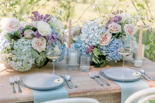 A close-up of a wedding table setting featuring blue and white floral centerpieces, glassware, and silverware on a rustic wooden table.