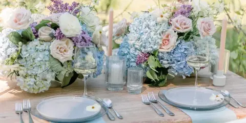 A close-up of a wedding table setting featuring blue and white floral centerpieces, glassware, and silverware on a rustic wooden table.