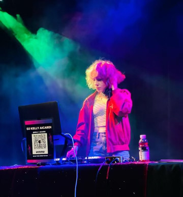 A DJ performing at an event with colorful stage lighting, standing behind a setup with a sign that reads 'DJ KELLY AICARDI'.