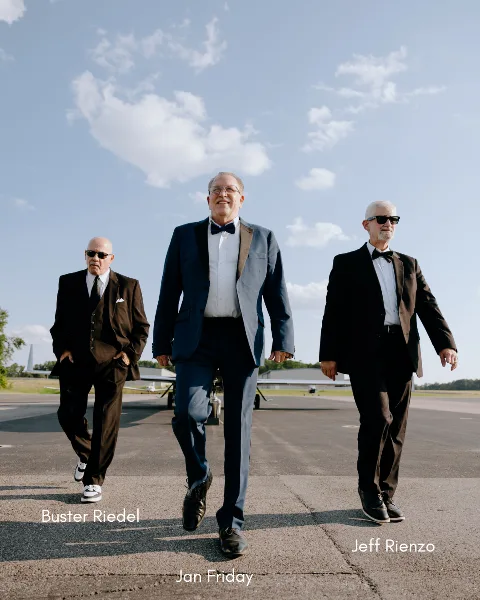 Three men in formal suits and bow ties walking on an airport tarmac, with the central figure labeled 'Jan Friday'.