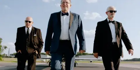 Three men in formal suits and bow ties walking on an airport tarmac, with the central figure labeled 'Jan Friday'.