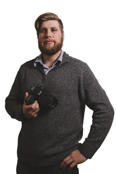A bearded man in a grey sweater holding a professional DSLR camera.