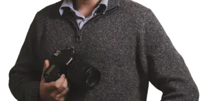 A bearded man in a grey sweater holding a professional DSLR camera.
