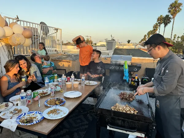 A chef in a grey uniform grilling meat and vegetables on a large flat-top hibachi grill for a group of people seated at a table on a rooftop.